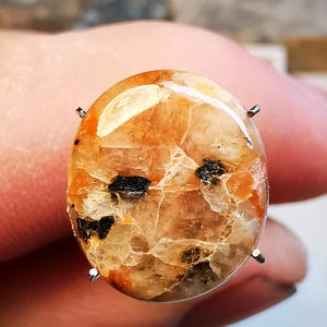 Puede incluir: Primer plano de una piedra preciosa tallada en cabujón de color naranja y blanco translúcido. La piedra tiene inclusiones negras y está engastada en un engaste de plata.
