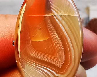 Pode incluir: Um cabochão de ágata polido de forma oval com um padrão em camadas de branco, laranja e amarelo. A pedra é translúcida, permitindo que a luz passe por ela.