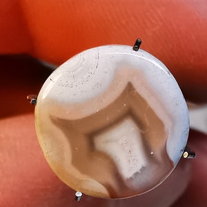 以下が含まれることがあります： 茶色とベージュの縞模様が入った丸い白いアゲートのcabochon。石は銀のベゼルにセットされ、4本の爪で固定されています。