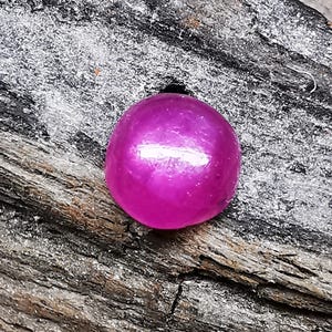 May include: A round, cabochon-cut gemstone with a vibrant, deep magenta-pink colour and a smooth, polished surface that reflects light. The stone features a translucent quality with subtle internal inclusions visible throughout its rounded dome shape.