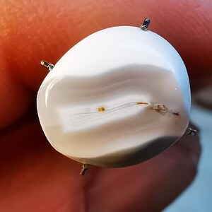 以下が含まれることがあります： 中心に薄い茶色の線が走る白いアゲートのcabochon。石は透明なプラスチック製の台座に4本の爪で留められています。