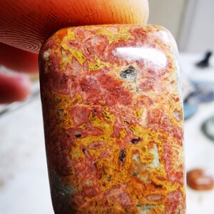 May include: A rectangular gemstone with rounded edges, displaying a vibrant mix of red, yellow, and orange hues. The stone's surface features intricate patterns and textures, creating a unique and natural appearance. The gemstone is held between two fingers.