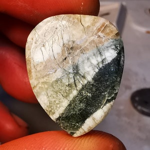 May include: A green and white gemstone with a unique pattern. The gemstone is shaped like a guitar pick.