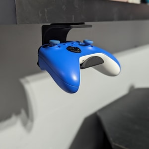 May include: A blue video game controller is hanging from a black shelf. The controller has white buttons and a black faceplate.
