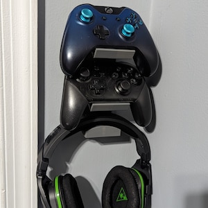 May include: Two black video game controllers with blue buttons are mounted on a wall with a black and green headset hanging below them.