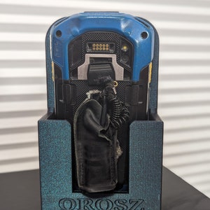 Puede incluir: Un escáner de mano azul y negro con una funda de cuero negro está en un soporte de impresión 3D azul turquesa con la palabra "OROSZ" impresa en la parte delantera.