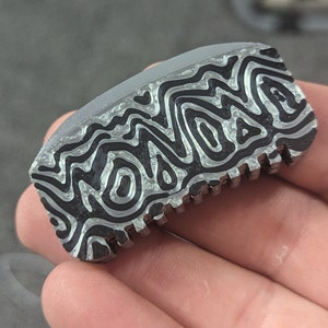 Op de afbeelding: Een close-up van een zilver en zwart gepatroneerd metalen object met een getextureerd oppervlak. Het object heeft een golvend, organisch ontwerp.