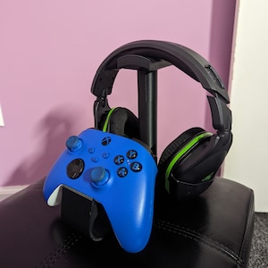 May include: A blue Xbox controller and black gaming headset with green accents are displayed on a black stand. The stand is on a black leather surface.