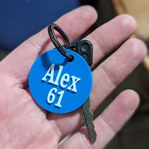 May include: A blue round keychain with the name "Alex 61" printed in white. The keychain is attached to a silver key.