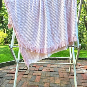 May include: A light pink blanket featuring a textured dimpled surface and a decorative fringe trim along the edges. The fabric has a consistent raised dot pattern across the entire surface, and the fringe consists of thin, hanging strands.