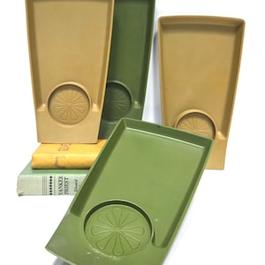 May include: Four rectangular trays in tan and olive green colors, each featuring a circular indentation with a molded flower petal design. The trays are arranged in a group, with two standing upright and two positioned in the foreground.