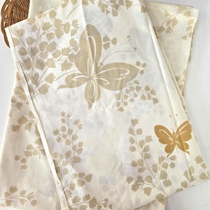 May include: Off-white fabric with a repeating pattern of tan butterflies and foliage. The butterflies are large and stylized, with detailed wing patterns. The fabric is folded, showing the pattern's repeat.