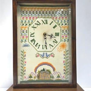 May include: A rectangular wooden clock with a glass front featuring a cross-stitch sampler face. The clock face displays green Roman numerals, black hands, and a colorful embroidered scene of a house, rainbow, sun, and floral patterns.