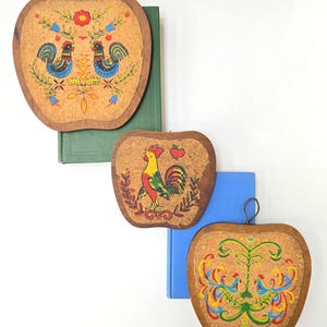 May include: Three apple-shaped cork trivets with wooden frames. Each trivet features a different colorful rooster design. The trivets are resting on top of green and blue books against a white background. The trivets have a metal loop for hanging.