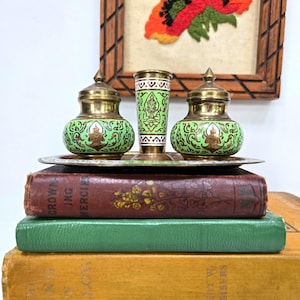May include: A set of three green and gold decorative containers with ornate designs, resting on a silver tray. The containers are placed on top of a stack of vintage books with brown, green, and yellow covers. An embroidered floral artwork hangs in the background.