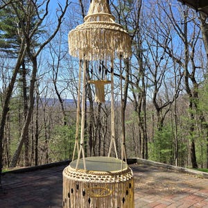 May include: A macrame hanging chair with a round, woven seat and a fringe border. The chair is suspended from a wooden ceiling and has a natural, cream color. The chair is hanging in front of a wooded area with a blue sky in the background.