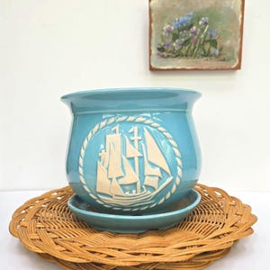 May include: A turquoise ceramic planter with a raised white sailboat design, set on a matching saucer and a round woven wicker mat. A small painting of flowers hangs in the background.