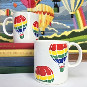 May include: Two white ceramic mugs with a hot air balloon design. The balloons are red, yellow, green, and blue. The mugs are sitting on a stack of books.