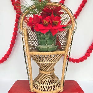 May include: A small, decorative peacock chair made of woven wicker, holding a green ceramic pot filled with red flowers. The chair sits on a red metal case, with red pom-pom garland in the background. The chair has a black and white trim.