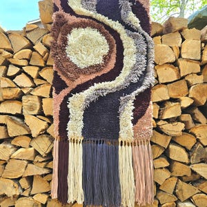 May include: A wall tapestry with a swirling abstract pattern in shades of brown, cream, purple, and gray. The tapestry has a fringe border.