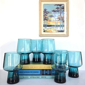 May include: A set of eight turquoise-colored drinking glasses with a dark base. The glasses are displayed on a stack of vintage books, with a framed landscape painting in the background. The glasses have a modern, geometric design.