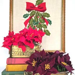 May include: A framed needlepoint featuring a red bow and holly. Below, two brass containers hold red and burgundy poinsettias. The containers sit atop a stack of vintage books, creating a festive holiday display.