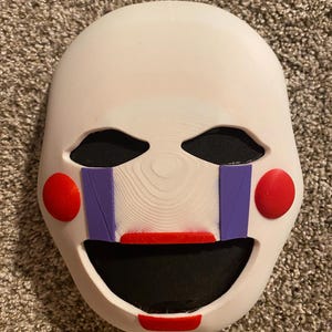 May include: A white mask with a smooth, rounded shape featuring black eye cutouts and a large black mouth opening. The mask includes two bright red circular cheek accents, two vertical purple stripes extending from the eyes, and red lip details.
