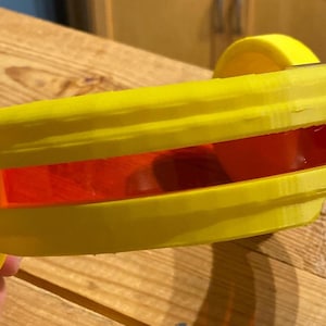 May include: A yellow, 3D-printed visor with a rectangular, red-tinted lens. The visor has a curved design and is held by a hand. The object is made of plastic and appears to be a prototype or a custom-made item.