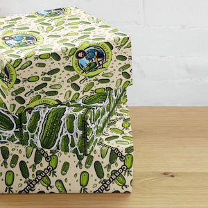 Peut inclure: Trois boîtes empilées avec un motif répété de cornichons verts sur un fond beige. Les boîtes présentent un graphisme circulaire avec une personne et le texte "Happy Little Pickles". Le mot "Félicitations" est imprimé sur les côtés.