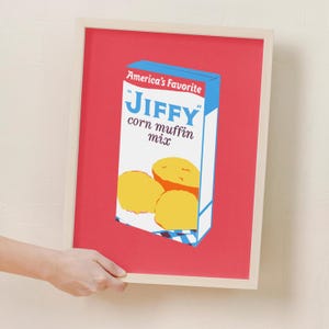Puede incluir: Una impresión enmarcada de una caja de mezcla para muffins de maíz Jiffy de estilo vintage. La caja es roja, blanca y azul con las palabras "America's Favorite" y "Jiffy corn muffin mix" en azul. Hay tres muffins de maíz amarillos en la caja.