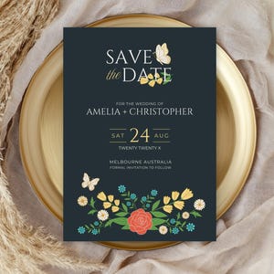 Op de afbeelding: Een donkerblauwe save-the-date kaart met gouden letters. De kaart heeft een bloemmotief met vlinders en de tekst "SAVE the DATE" voor de bruiloft van Amelia en Christopher op zaterdag 24 augustus 2020 in Melbourne, Australië.