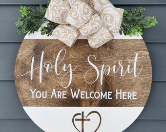 May include: A round wooden sign with the words "Holy Spirit You Are Welcome Here" in white script. The sign has a brown wood grain top and a white bottom section. It is decorated with a beige bow and greenery.