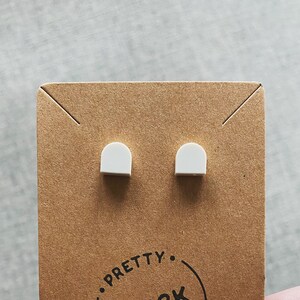May include: A pair of white, arch-shaped stud earrings on a brown card. The card has the text "Pretty Brick Ears" in a circular design. The earrings are small and minimalist.