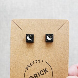 May include: A pair of black square stud earrings with a white crescent moon and gold stars design. The earrings are displayed on a brown card with the text "Pretty Brick Ears".