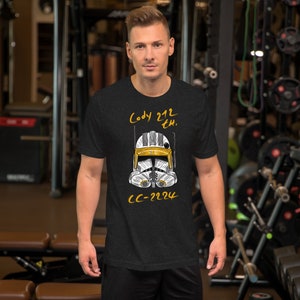 May include: A black t-shirt featuring a white and yellow illustration of a Star Wars clone trooper helmet. The text "Cody 212th" and "CC-2224" is printed on the shirt.