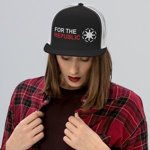 Op de afbeelding: Een zwart-witte truckercap met een wit mesh achterpaneel. De cap heeft de tekst "FOR THE REPUBLIC" in rood en een wit symbool aan de voorkant.