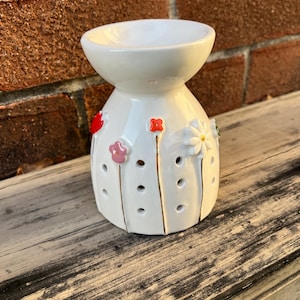 May include: A white ceramic essential oil diffuser with a flower design. The diffuser has a bowl-shaped top and a cylindrical base with holes. The flowers are red, pink, and white with gold stems. The diffuser sits on a wooden surface.