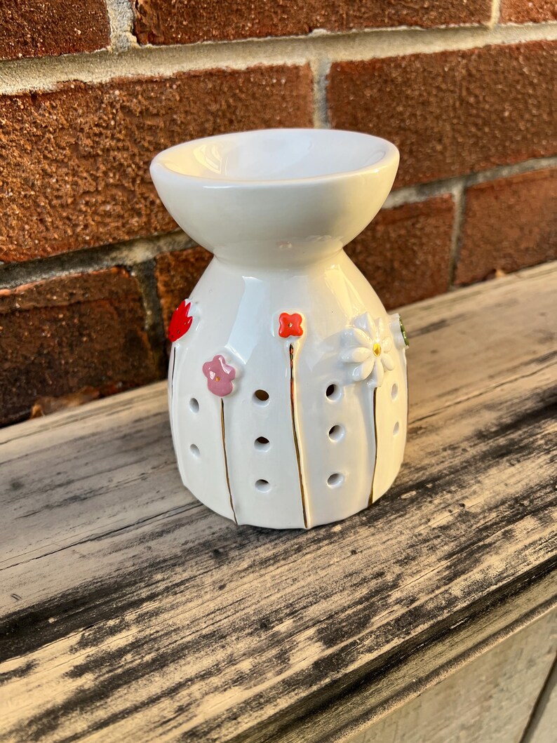 May include: A white ceramic essential oil diffuser with a flower design. The diffuser has a bowl-shaped top and a cylindrical base with holes. The flowers are red, pink, and white with gold stems. The diffuser sits on a wooden surface.