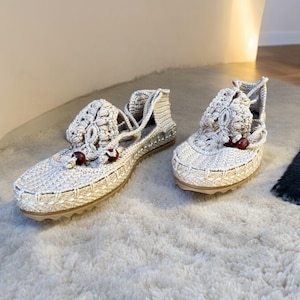 May include: A pair of beige crocheted sandals with red bead accents. The sandals have a lace-up design and are made of a soft, lightweight material.