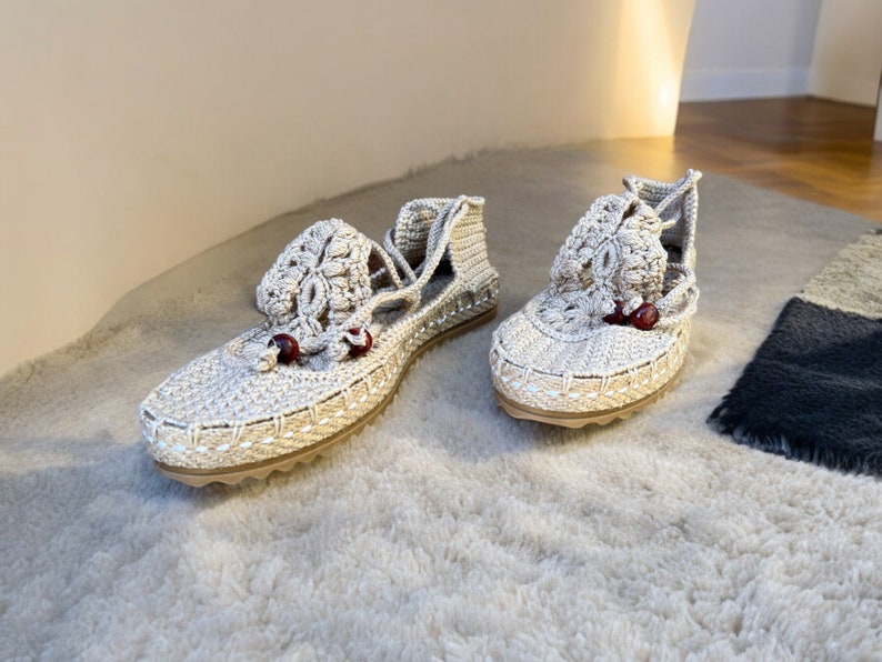 May include: A pair of beige crocheted sandals with red bead accents. The sandals have a lace-up design and are made of a soft, lightweight material.
