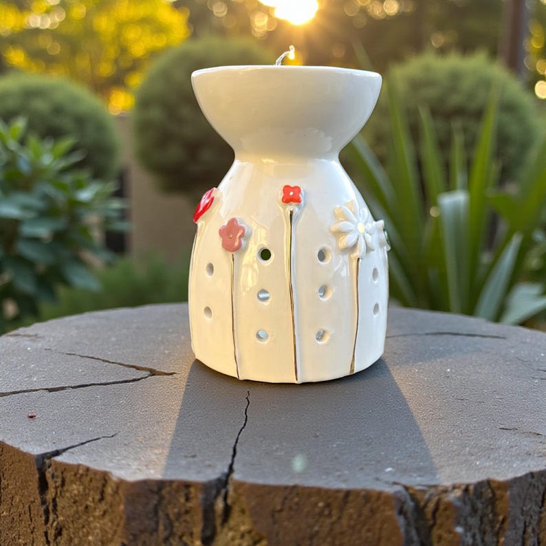 May include: A white ceramic essential oil diffuser with a floral design. The diffuser has a wide top and a bulbous base, decorated with red and pink flowers and small holes. A lit candle sits inside.