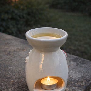 May include: A white ceramic essential oil diffuser with a lit tea light candle inside. The diffuser has a round top for holding oil and a decorative design. The background is blurred, showing greenery and a soft, natural light.