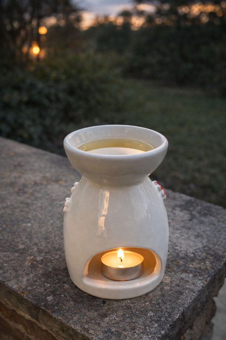 May include: A white ceramic essential oil diffuser with a lit tea light candle inside. The diffuser has a round top for holding oil and a decorative design. The background is blurred, showing greenery and a soft, natural light.