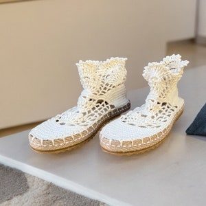 May include: A pair of white crocheted espadrille shoes with a lace-like design. The shoes have a flat sole and a closed toe.
