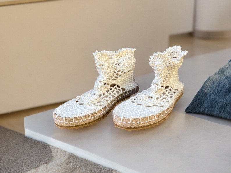 May include: A pair of white crocheted espadrille shoes with a lace-like design. The shoes have a flat sole and a closed toe.