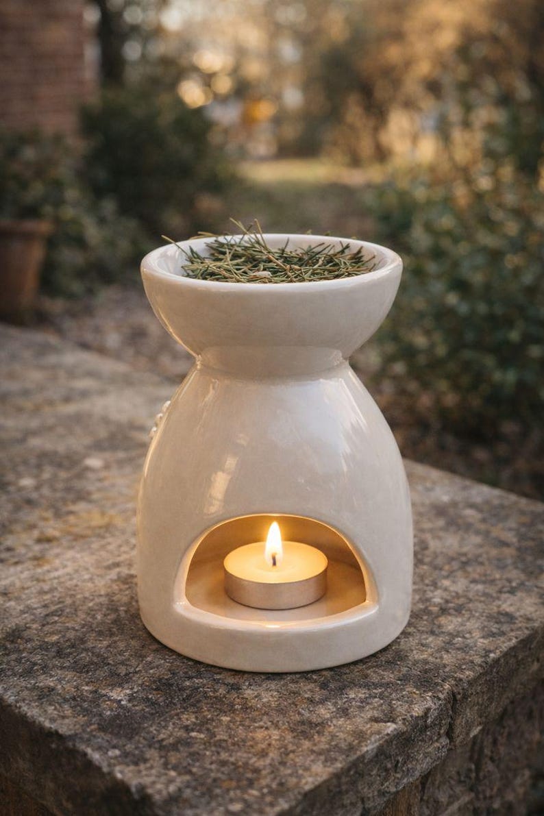 May include: A cream-colored ceramic essential oil diffuser with a lit tea light candle inside. The top bowl holds sprigs of greenery. The diffuser has a rounded shape with a wide base and a narrower top, creating a decorative and functional item.