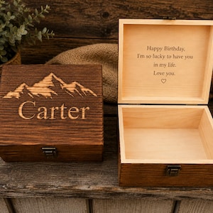 May include: Two wooden boxes with a dark brown finish. One box is closed and features a laser-engraved mountain range and the name Carter. The other box is open, revealing a light wood interior with the text: Happy Birthday, I'm so lucky to have you in my life. Love you. with a heart symbol.