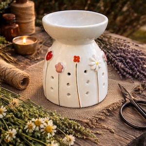 May include: A white ceramic essential oil diffuser with floral accents. The diffuser has a wide top and a rounded base, decorated with red, pink, and white flowers. It sits on a wooden surface with other aromatherapy items.