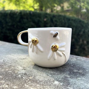 May include: White ceramic mug with gold accents, featuring daisy and bee embellishments. The mug has a gold handle and gold detailing on the daisy centres. The mug is sitting on a grey stone surface.