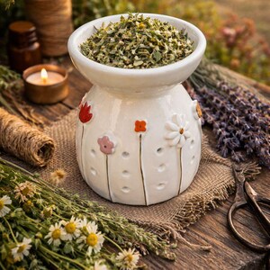 May include: A white ceramic essential oil diffuser with floral accents. The diffuser is filled with dried herbs and sits on a wooden surface. Other items include dried flowers, a candle, and twine.